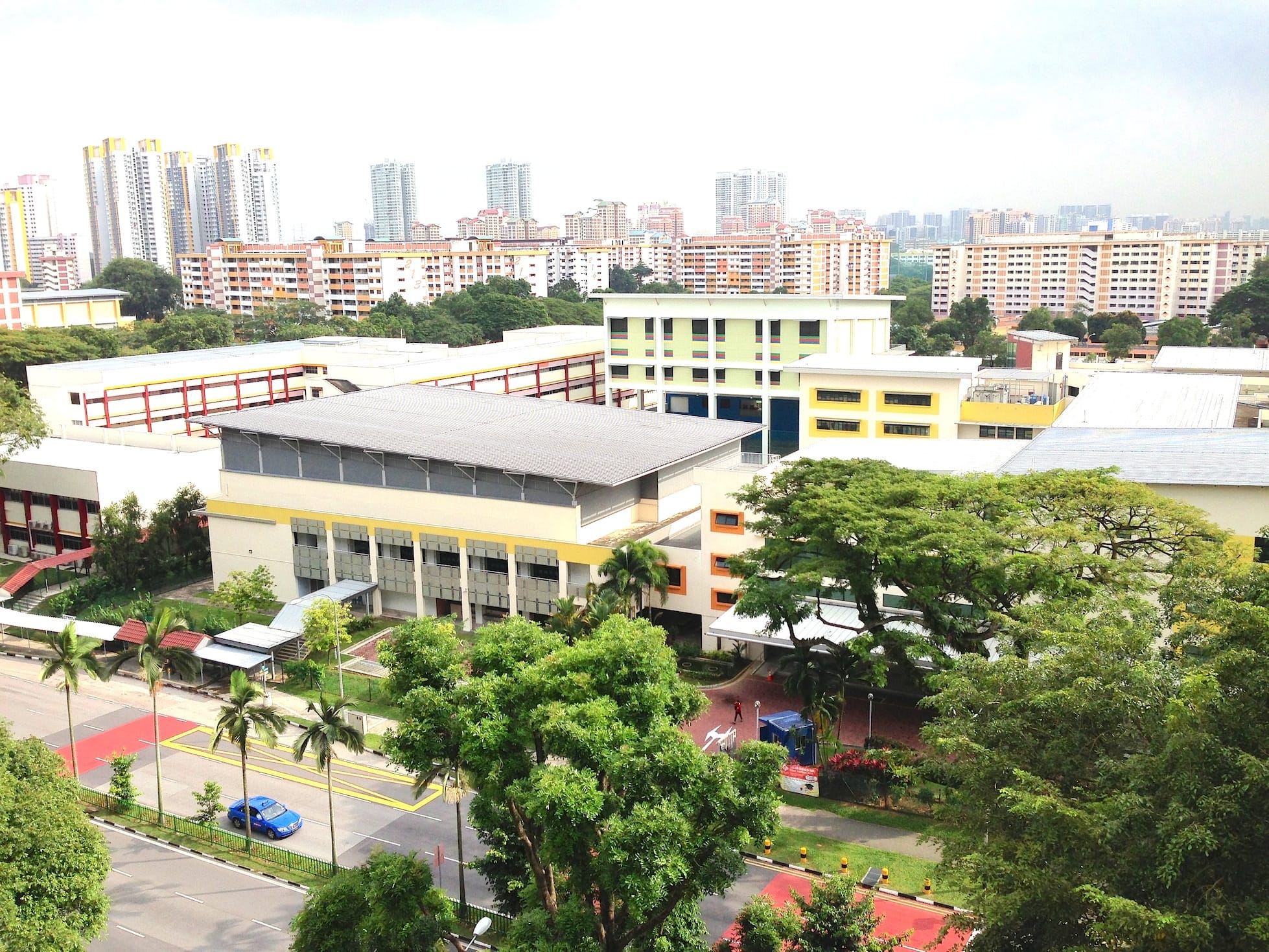 Aerial view of a school campus with colorful buildings, trees, a road with a blue car, and tall residential blocks behind.